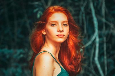 A woman with vibrant red hair standing in a forest setting.