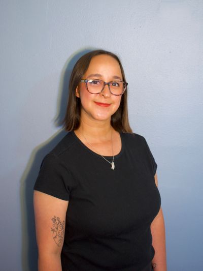 Woman with glasses and tattoo wearing a black shirt against a blue wall.