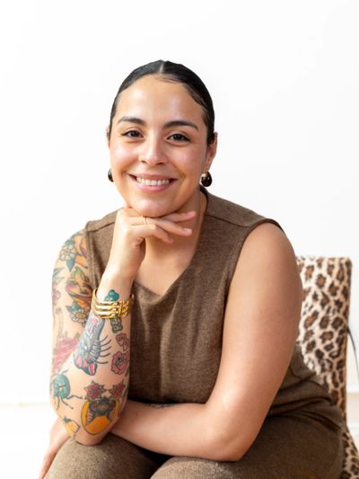 Smiling woman with tattoos and gold jewelry, wearing a brown sleeveless top.