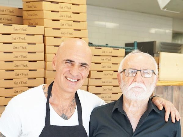 Two men smiling in a pizzeria with stacked pizza boxes behind them.
