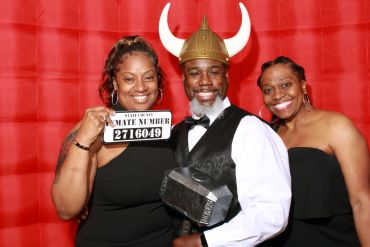 Three people posing in front of a red backdrop, one holding a sign and another wearing a Viking helmet.