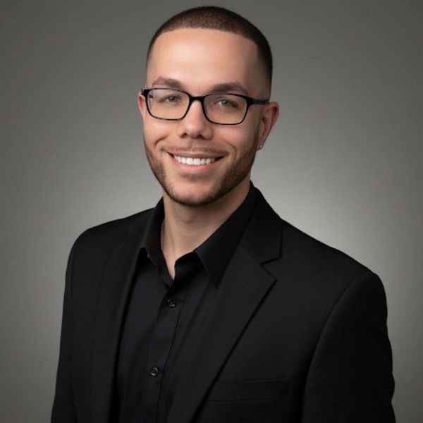Smiling man in black suit and glasses against gray background.