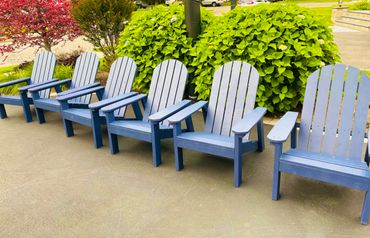 Row of blue Adirondack chairs on a patio with greenery behind.