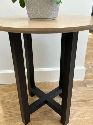 Round wooden table with black crossed legs and a plant on top.