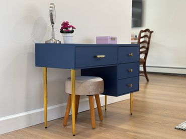 Blue vanity desk with gold legs and a cushioned stool in a stylish room.