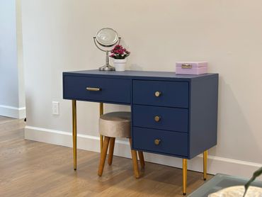 Navy blue vanity desk with gold legs and a small stool in a minimalist room.