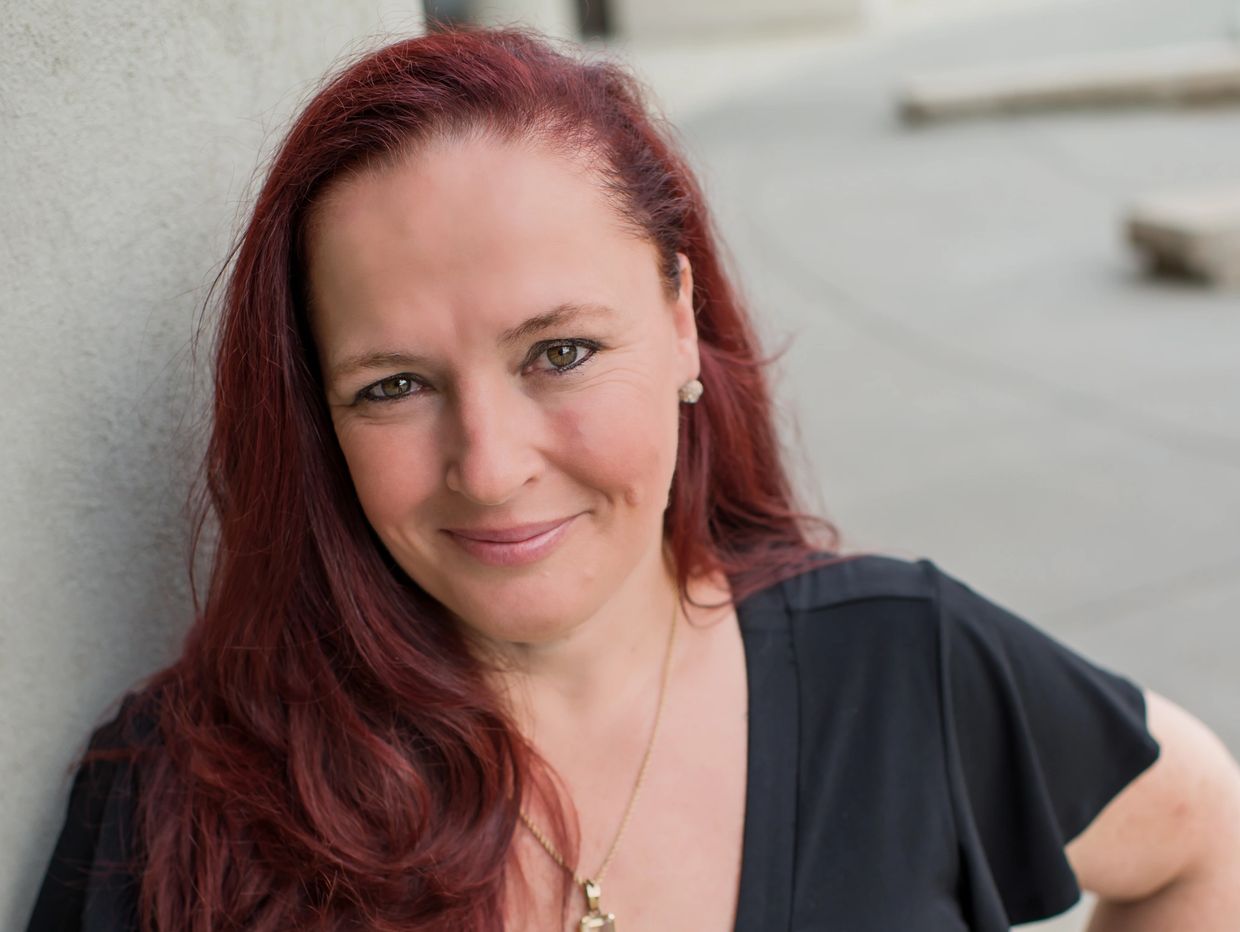 Smiling woman with red hair and a black top leaning against a wall.