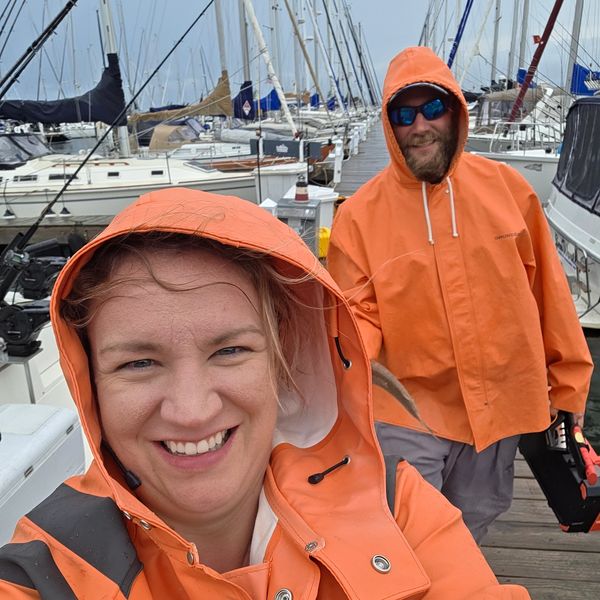 Two people in orange rain jackets smiling at a marina dock with boats.