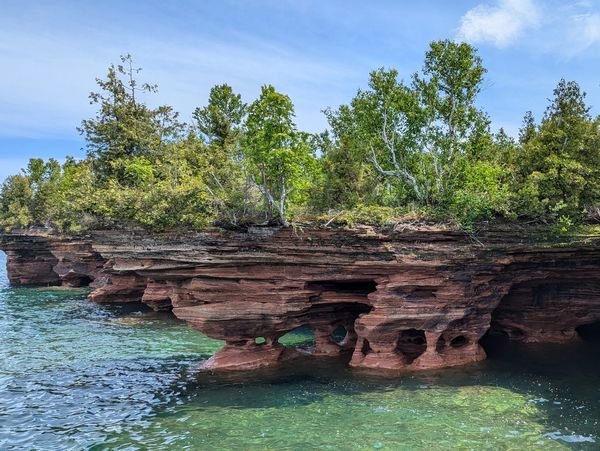 Rocky cliffs with caves by clear green water and trees above.