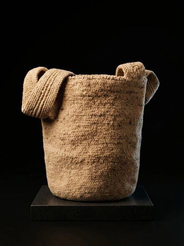 A textured woven basket with a folded strap on a black pedestal.