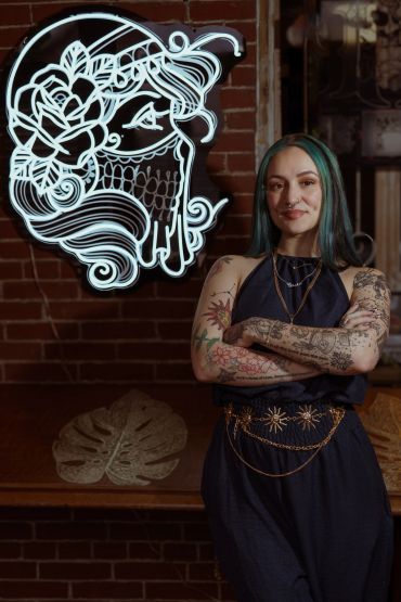 Tattooed woman with crossed arms in front of a neon art piece.