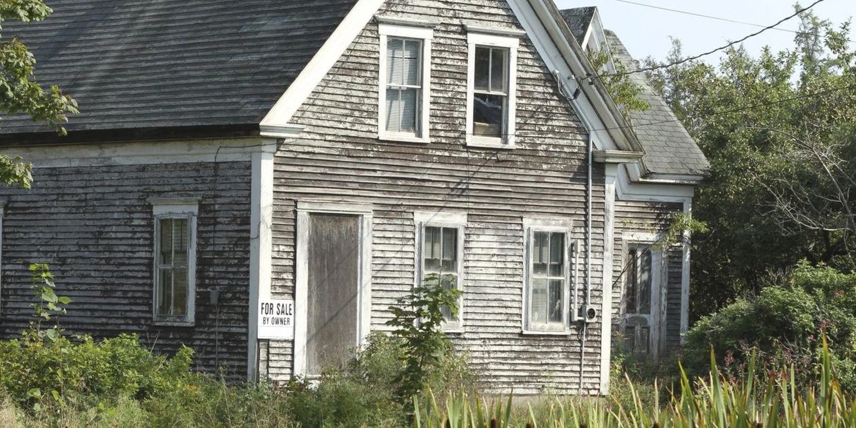 Old weathered house with peeling paint and "For Sale By Owner" sign overgrown by plants.