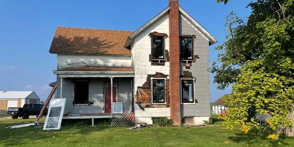 Old two-story house with boarded windows and a chimney in a grassy yard.