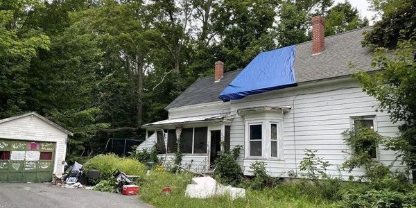 Old white house with a blue tarp on the roof surrounded by overgrown grass and clutter.