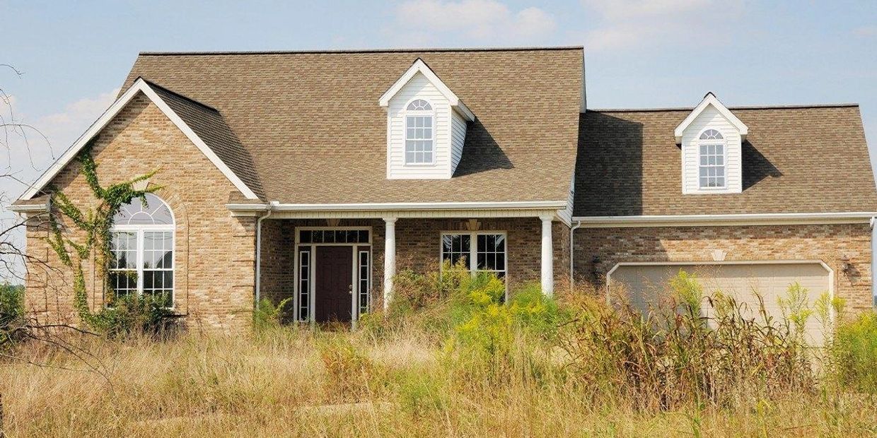 A brick house with overgrown grass and plants in front.