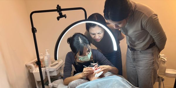 A beautician applies eyelash extensions while two women watch closely.