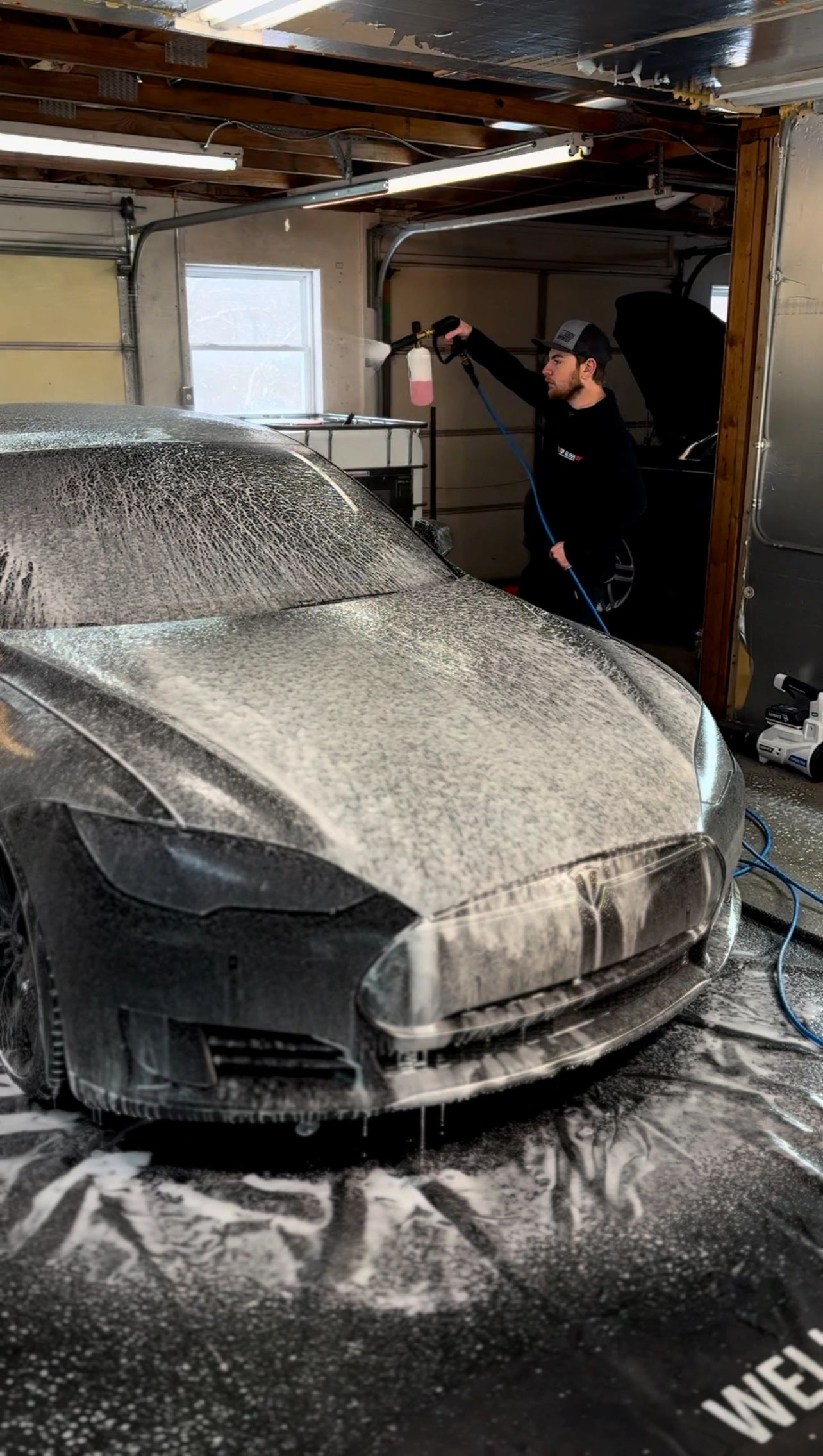 A man using foam cannon to wash a black car in a garage.