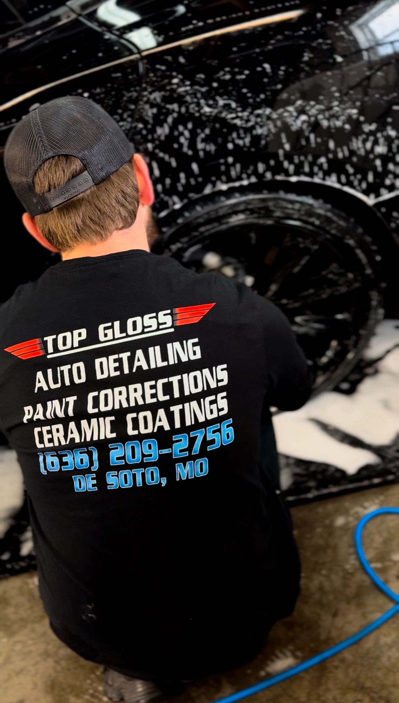 A person wearing a Top Gloss shirt washing a black car covered in soap.