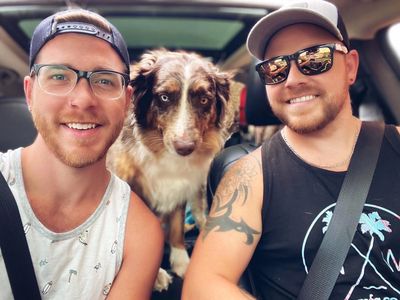 Two men and a dog smiling inside a car, all wearing seatbelts.
