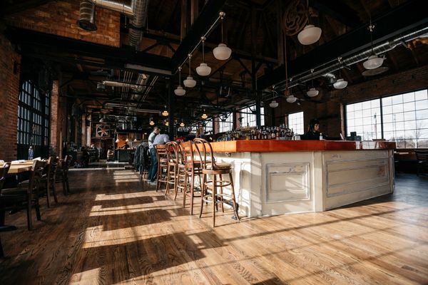 Spacious industrial-style restaurant interior with large windows, wood floors, & a central bar area.