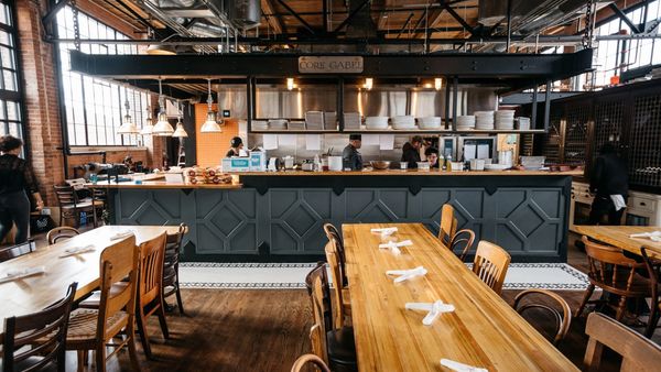 Industrial-style restaurant with open kitchen, wood tables, & a modern counter in a bright space.