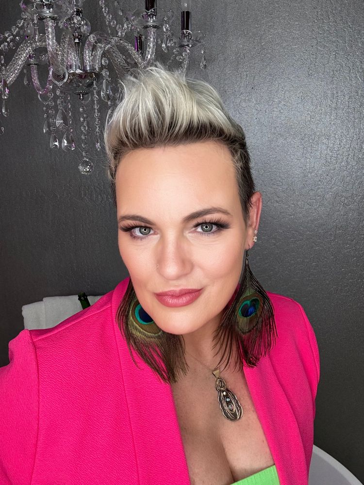 A photo of Kimber wearing a hot pink blazer jacket with peacock earrings and a metal vulva necklace.