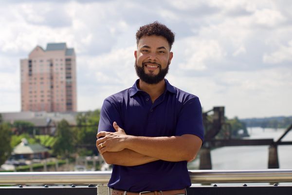 Smiling man with crossed arms standing outdoors with a city and river in the background.