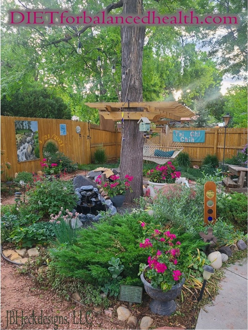 A backyard landscape with flowering plants, trees, fountain and hammock.