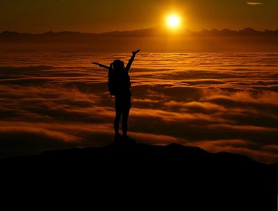 Silhouette of a person celebrating atop a mountain during a golden sunset.