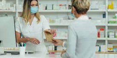 A female pharmacist wearing a blue face mask hands a small brown paper bag to a customer across a ph