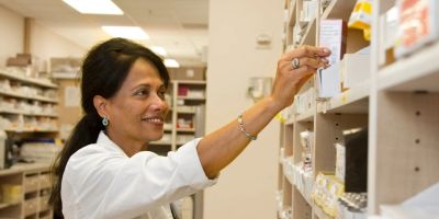 A pharmacist in a white coat reaches for a medication box on a pharmacy shelf.