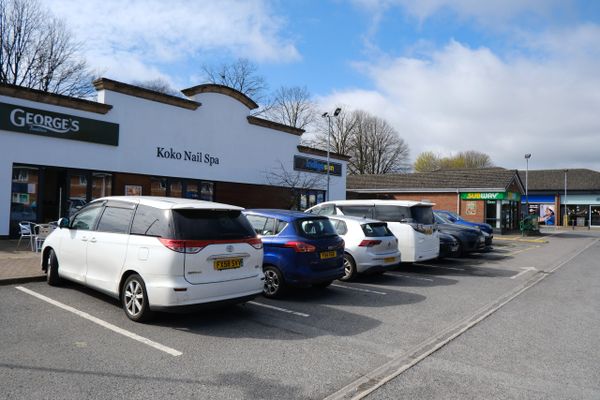 Parked cars in front of shops including Koko Nail Spa and Subway on a clear day.