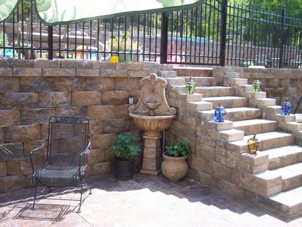 Outdoor stone patio with wrought iron chair, decorative fountain, potted plants, and stone stairs adorned with lanterns.