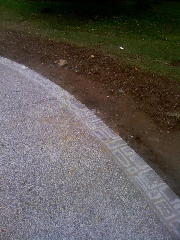Curved concrete pathway with stone border next to soil and grass.