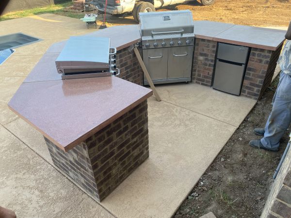 Outdoor kitchen setup with grill, fridge, and countertop on brick base.