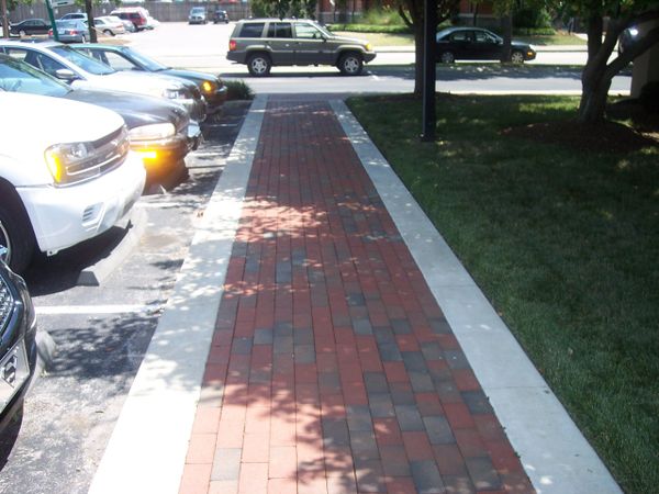 A brick sidewalk bordered by concrete with parked cars and a grassy area beside it.