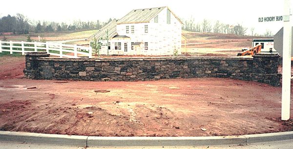 Empty lot with stone wall and house in the background on a rural street.