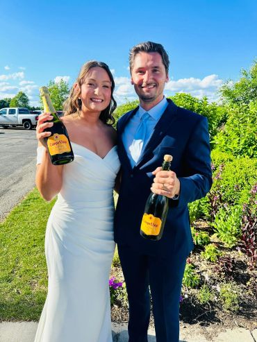 Newlywed couple smiling with champagne bottles outdoors.