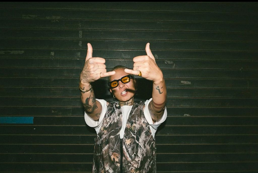 Person with tattoos and orange glasses showing middle fingers in front of a dark wall.