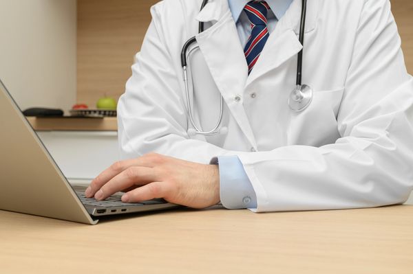 Doctor in a lab coat with a stethoscope sitting at a desk in front of a laptop