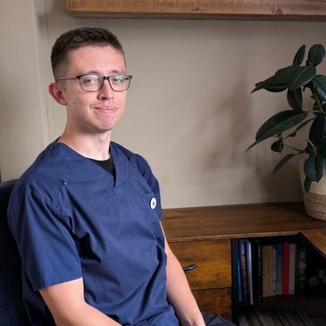 Young man in navy scrubs sitting in a cozy office space with books and a plant.