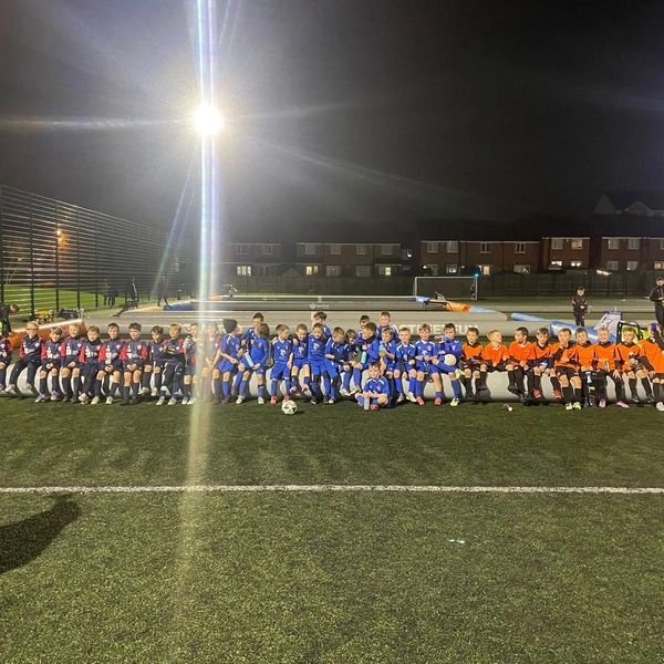 Three youth soccer teams in different uniforms sit side by side on a bench at night.