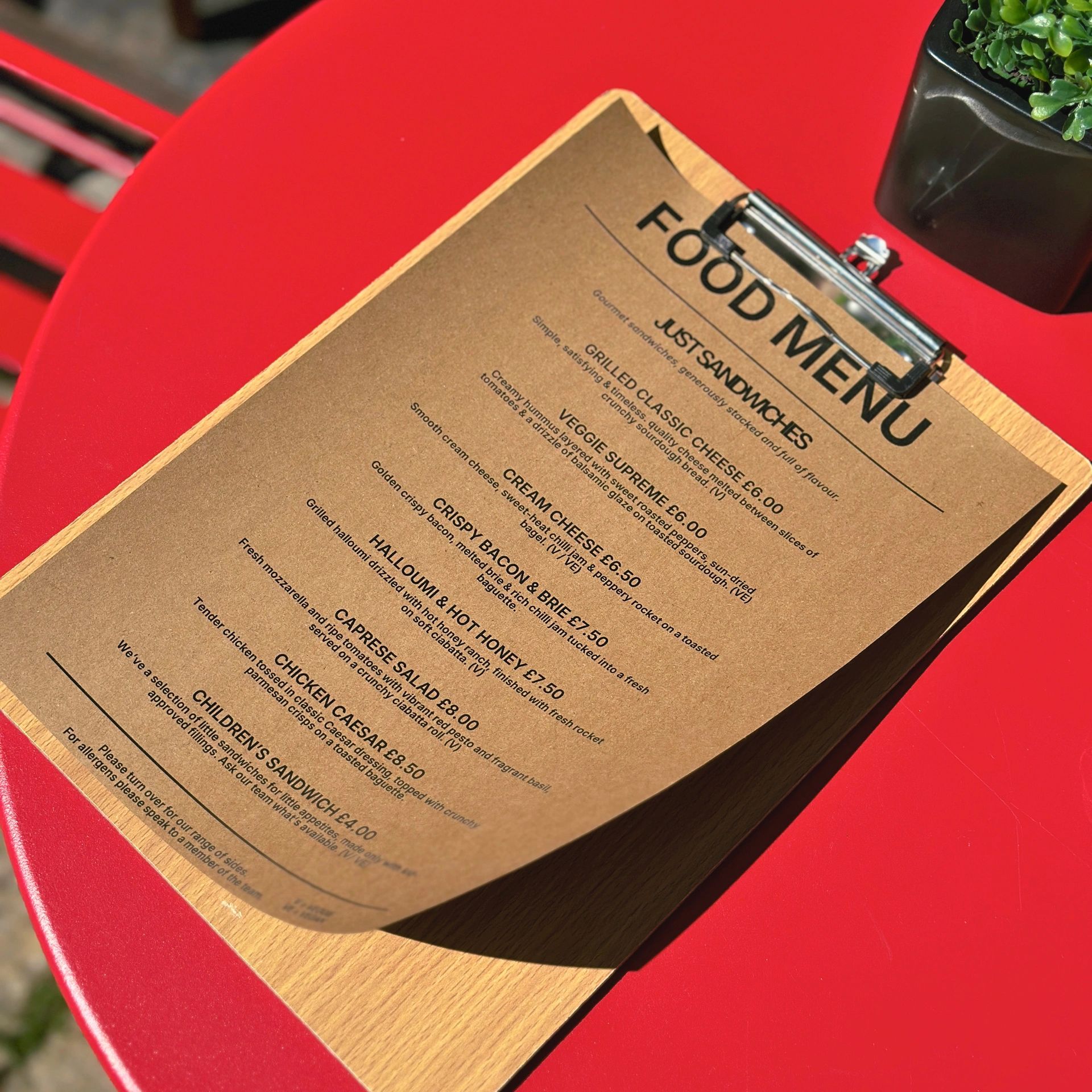A food menu on a wooden clipboard placed on a vibrant red table with a small plant.