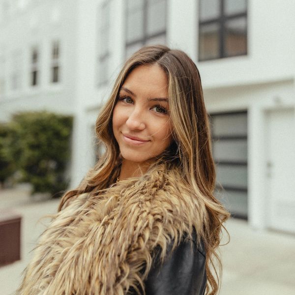 Young woman with long hair wearing a fur-lined jacket, smiling outdoors.
