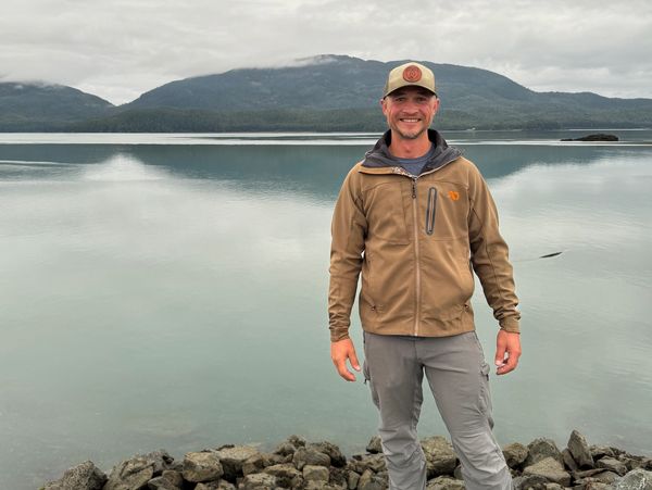 Man smiling by a rocky lakeshore with mountains and cloudy sky.