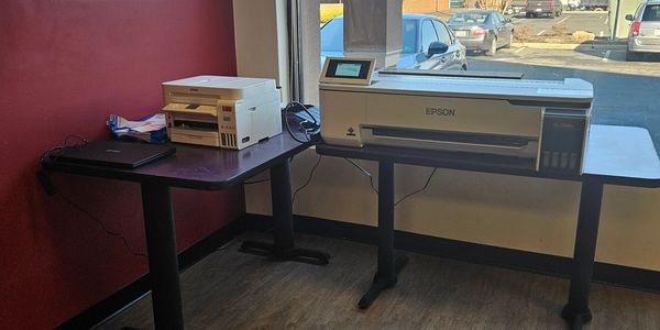 Two Epson printers on adjacent tables near a window with a parking lot view.