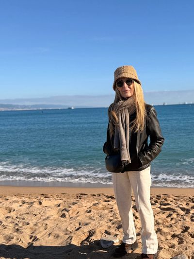 Woman in a hat and scarf stands on a sunny beach with blue sky and ocean.