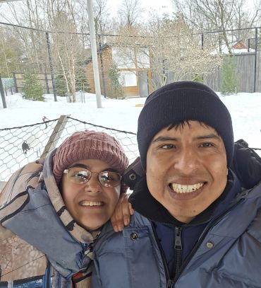 A smiling couple bundled up in winter clothes outdoors in a snowy setting.