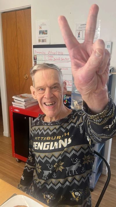Elderly man wearing a Pittsburgh Penguins sweater shows a peace sign and smiles.