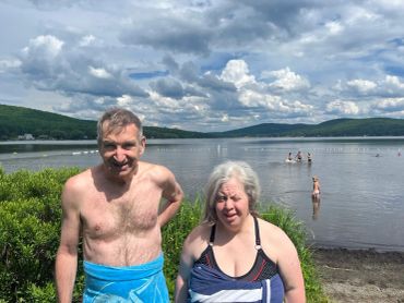 Two people smiling by a lakeside on a partly cloudy day.